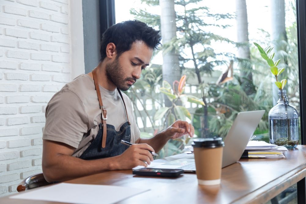 Man working at a cafe table with a laptop, notebook, and coffee by a bright window