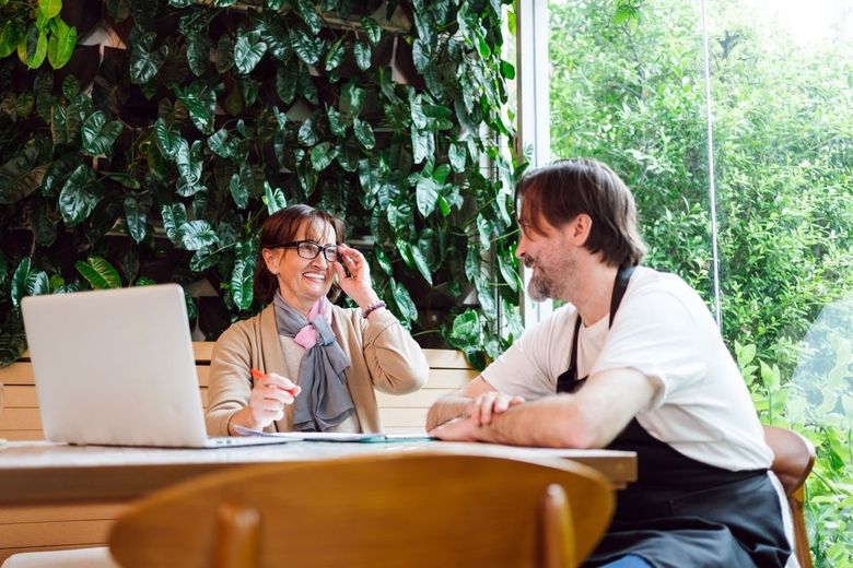 Two people talking at a table with a laptop in a bright, leafy office setting