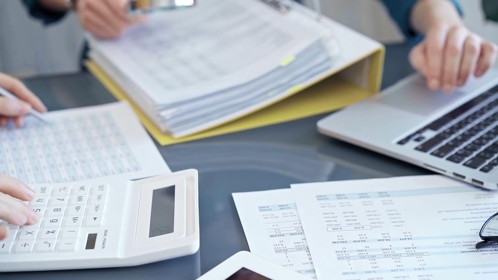 Hands using calculator and laptop on desk with paperwork and charts