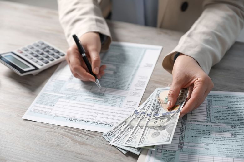 Hands filling out tax forms beside a calculator and cash on a desk
