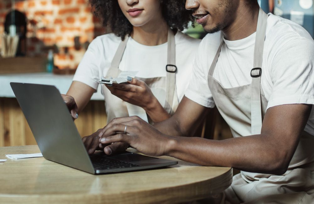 Two people in aprons using a laptop and smartphone at a café table