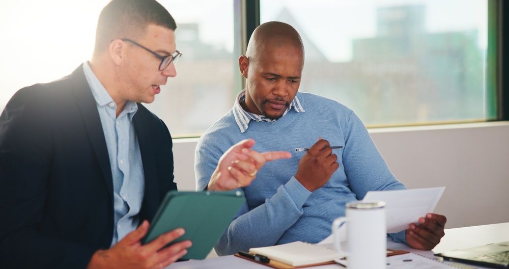 Two coworkers reviewing documents at a table, one holding a tablet and the other reading a phone.
