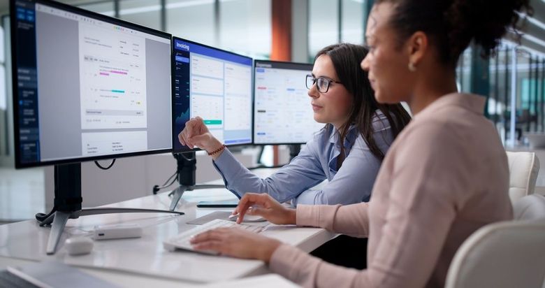 Two coworkers discussing data on dual monitors in a modern office
