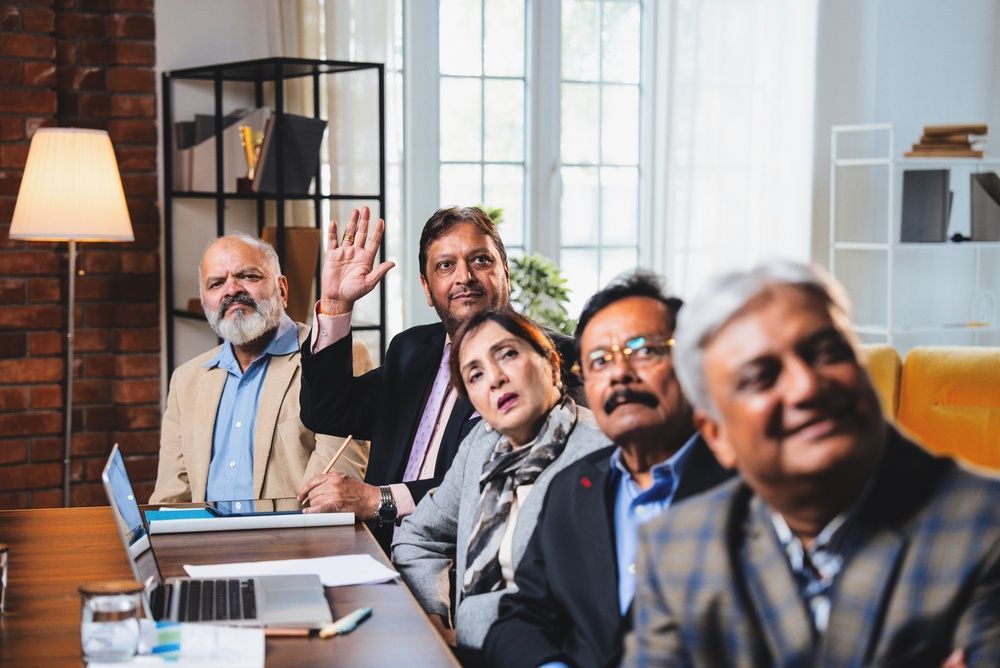 Business meeting with five people seated around a table in a bright office, one man raising his hand.