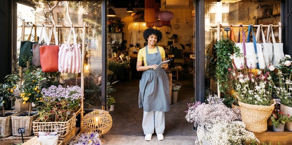 Shopkeeper standing in a flower-filled storefront, holding a tray among hanging clothes and potted blooms