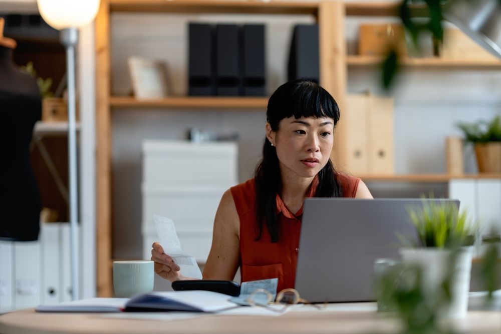 Woman working at a desk with a laptop, holding papers in a bright office.