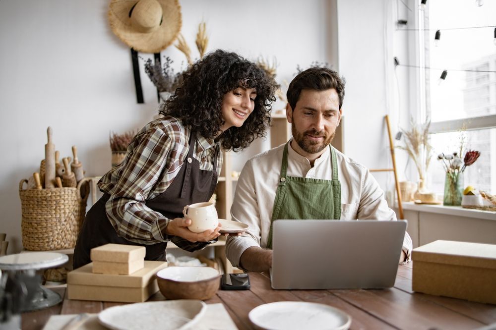 Two people working on a laptop at a kitchen table with boxes and wrapped goods nearby