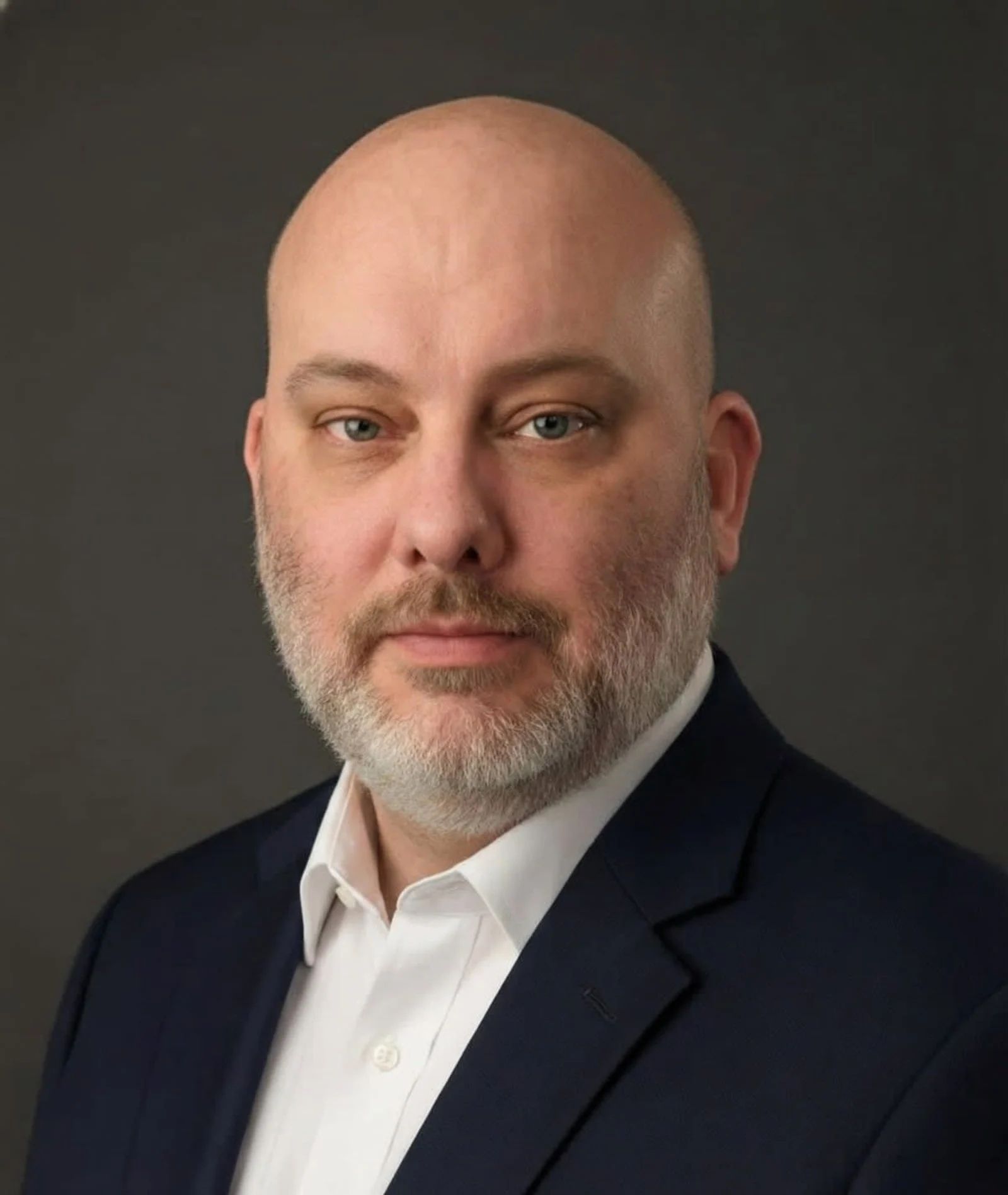 Studio portrait of a bald man with a gray beard in a dark suit and white shirt against a dark background