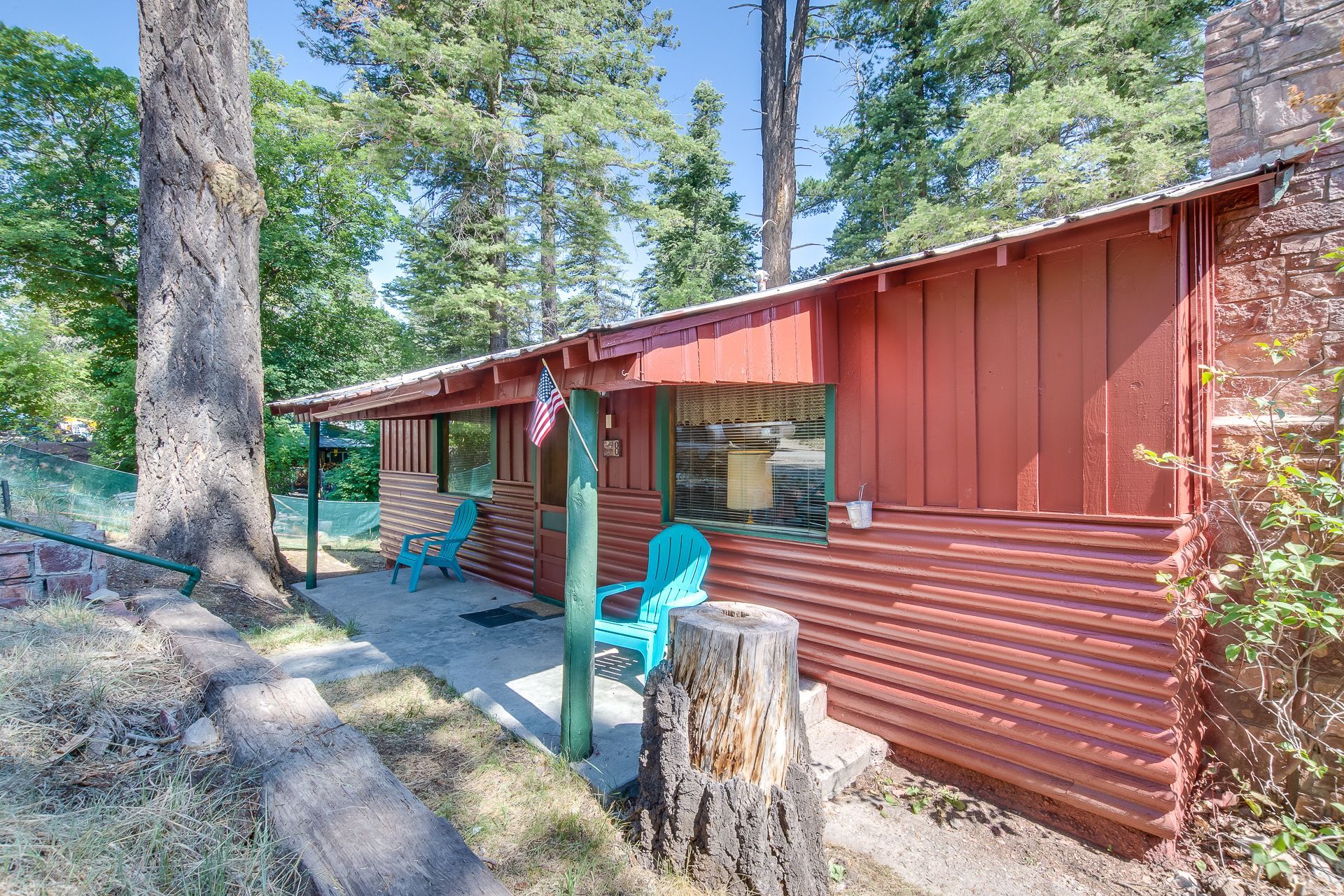A small, red-sided cabin nestled among pine trees, featuring a shaded front patio with two turquoise chairs.