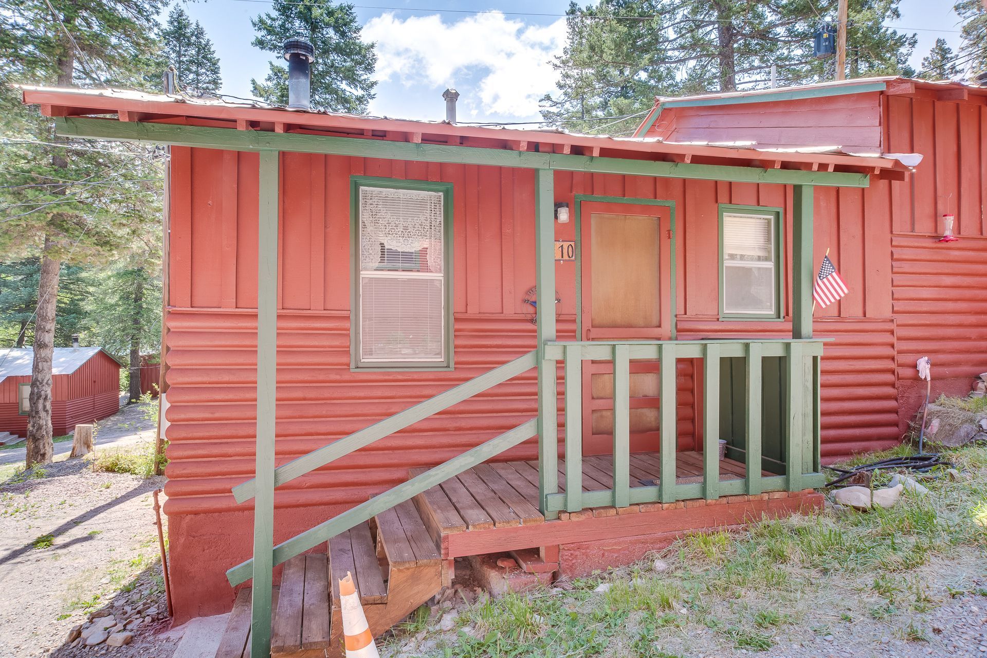 A rustic, red-sided cabin with a green-trimmed front porch and stairs nestled in a wooded area.