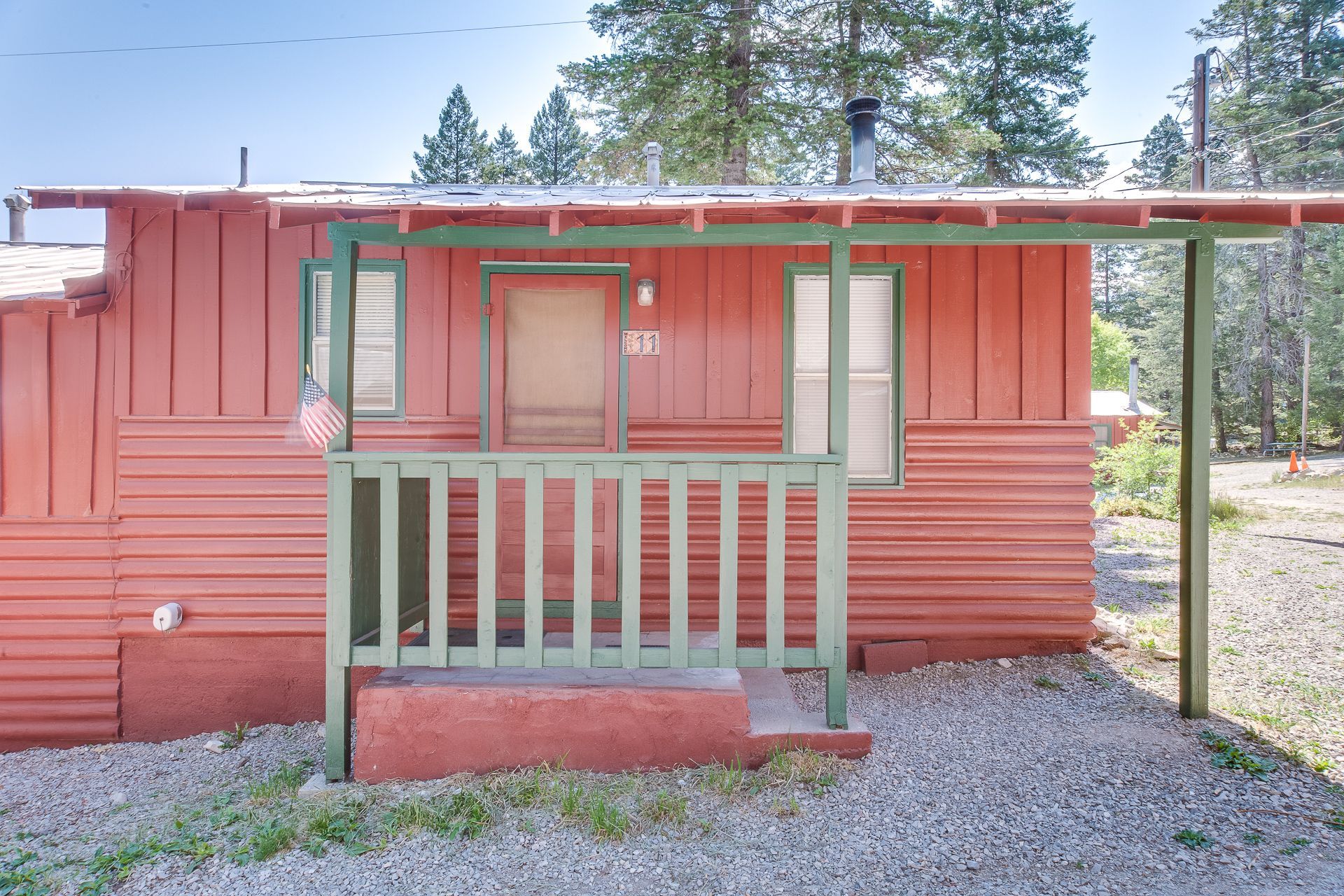 A small red wooden cabin with green trim, a front porch, and a gravel yard surrounded by pine trees.