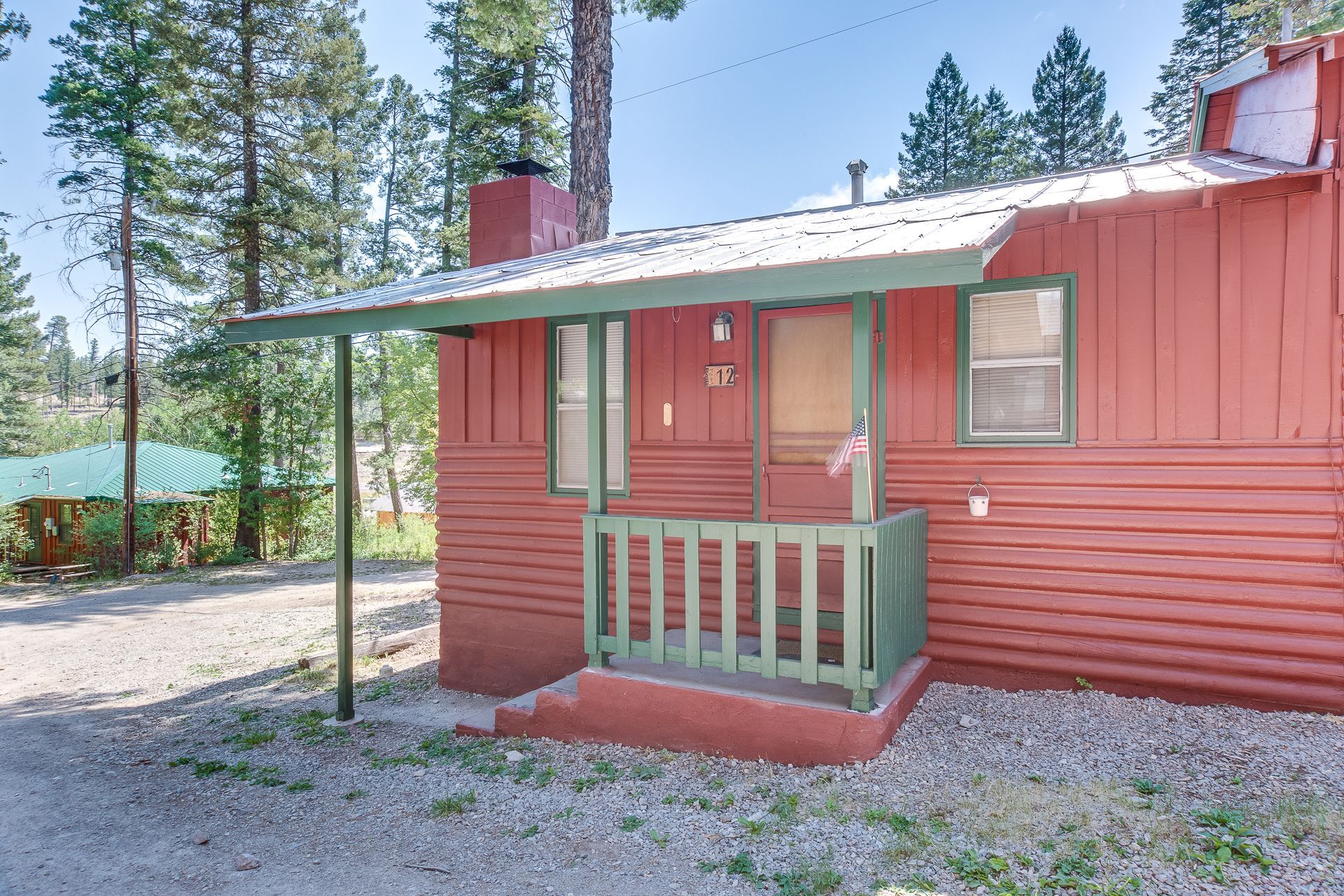 A small red wooden cabin with a green-railed porch and metal roof, situated in a wooded area with gravel ground.