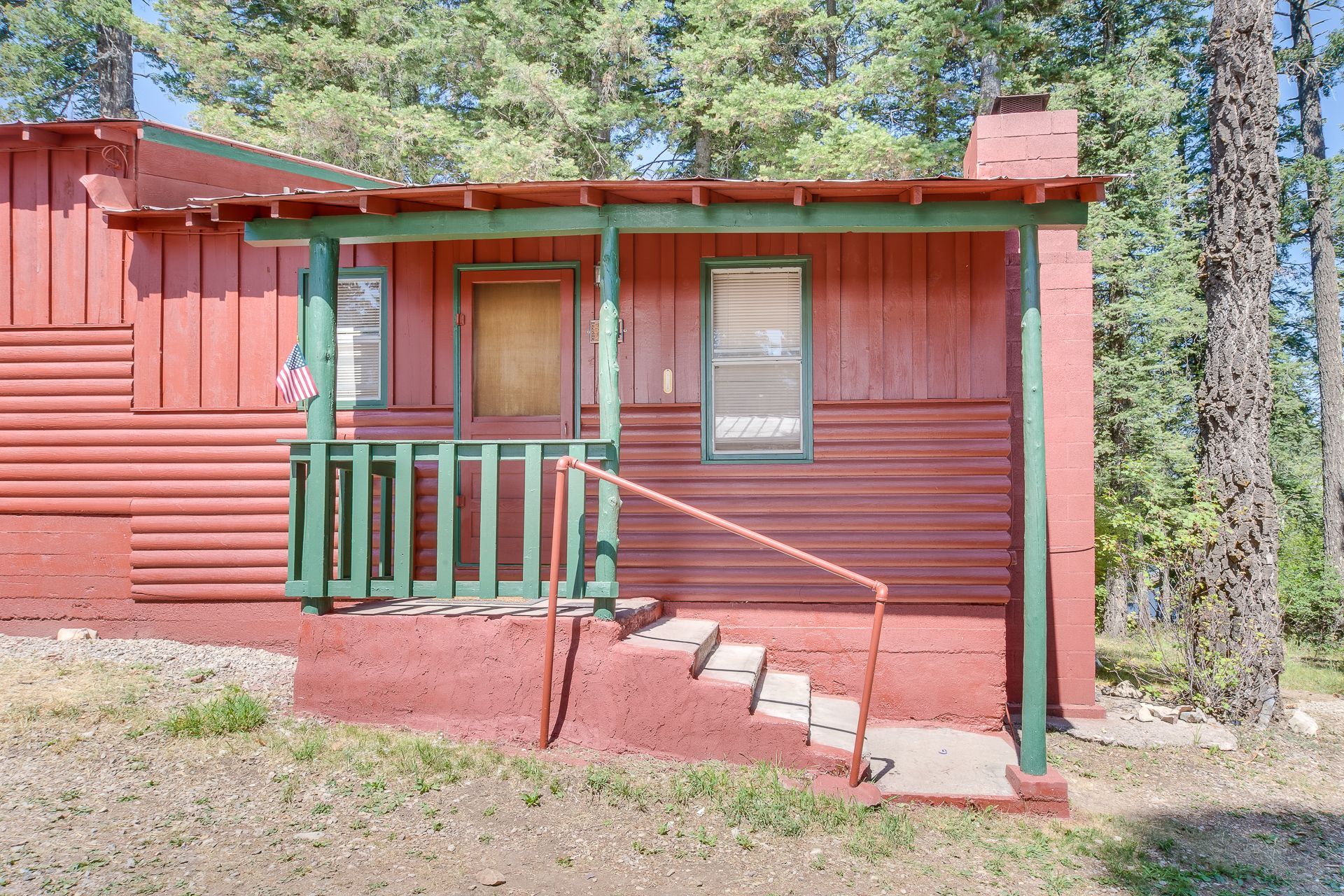 A small red cabin nestled among trees features a porch with green railings, a central door, and concrete steps.