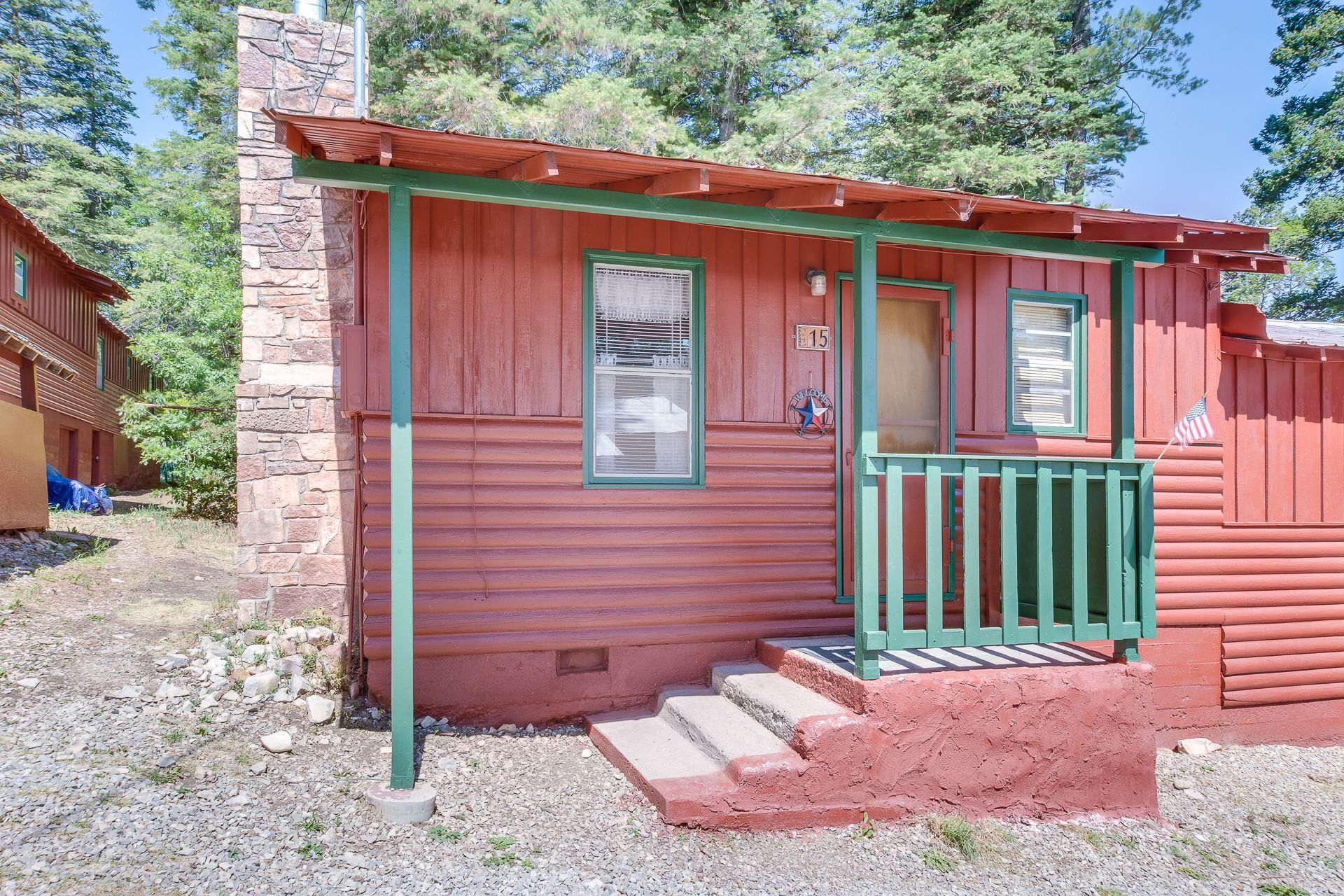 A small, red wooden cabin with a stone chimney, green porch railings, and steps, surrounded by trees on a gravel lot.