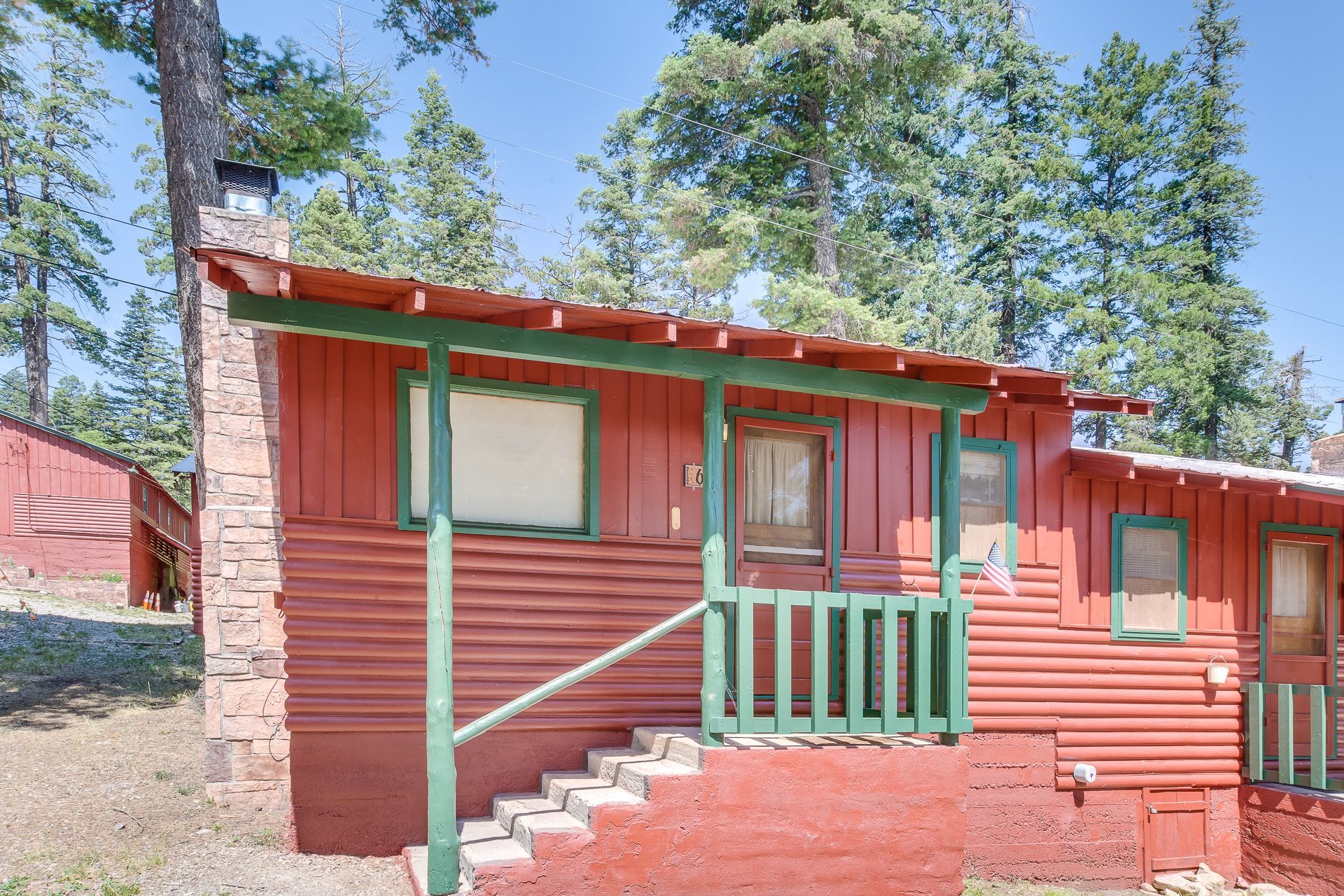 A red wooden cabin with a green-trimmed porch and stone chimney, nestled among tall pine trees on a sunny day.