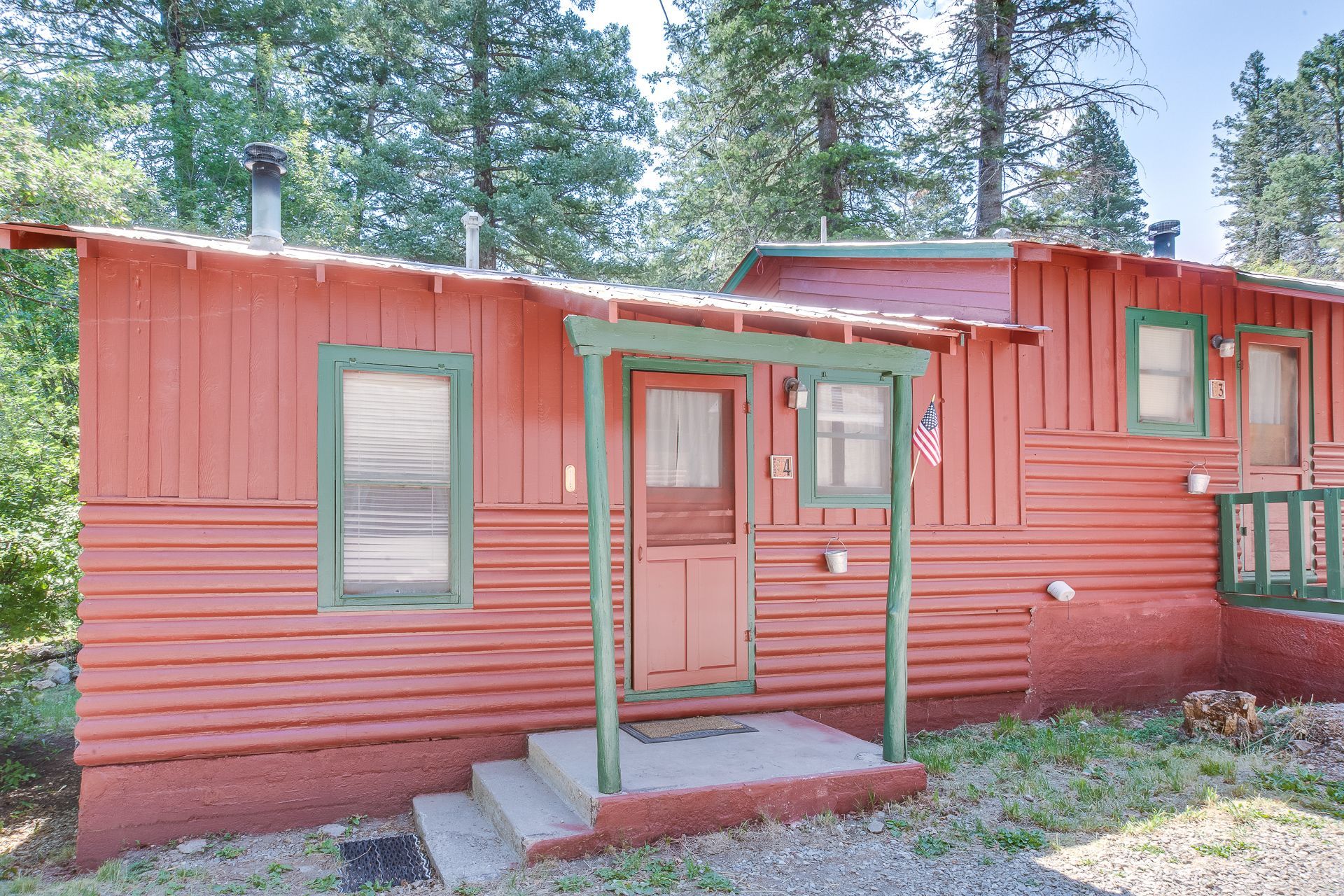 A rustic red cabin with green trim, a small front porch, and a concrete stoop, nestled among tall evergreen trees.