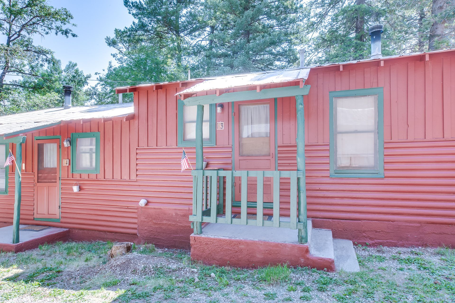 Two small, rustic cabins with red corrugated siding and green trim sit in a wooded area.