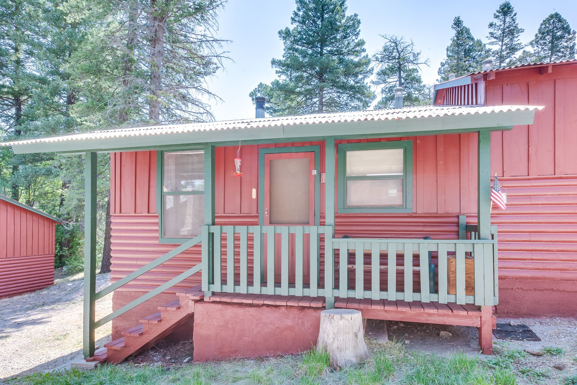 A red cabin with a covered front porch, wooden railings, and green trim set among evergreen trees.