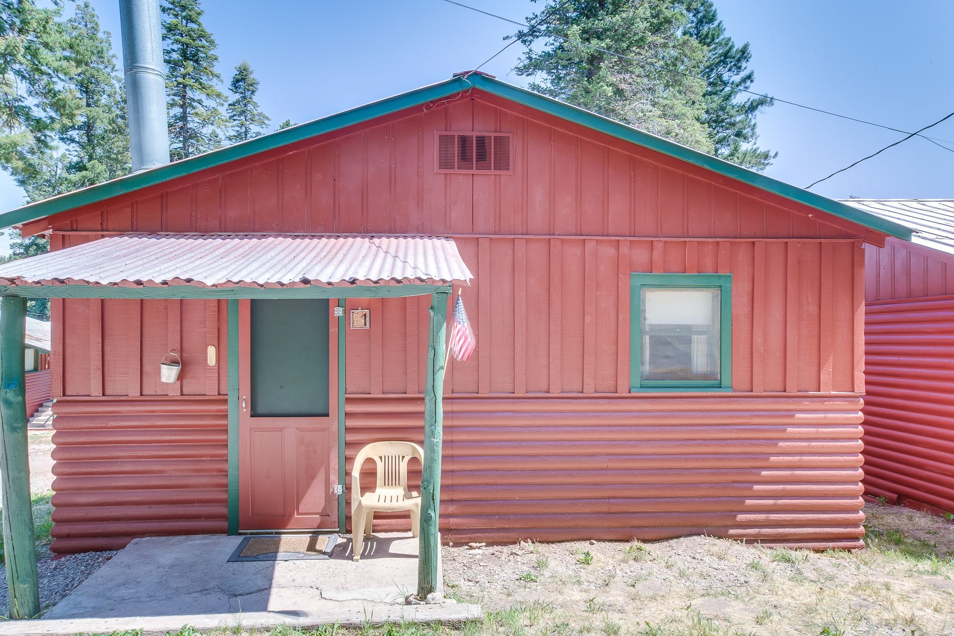 A small red cabin with horizontal wood siding, a green-trimmed window, and a front porch with a white plastic chair.