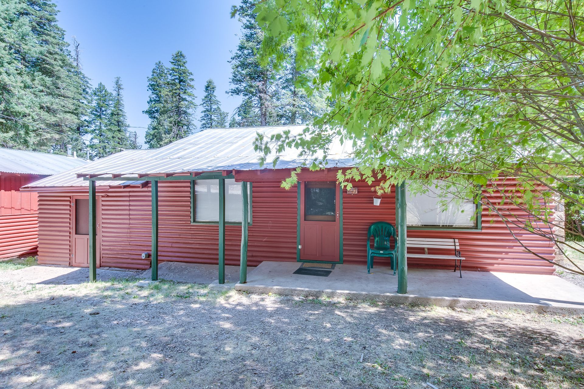 A red, single-story cabin with a metal roof and front porch, surrounded by trees on a sunny day.