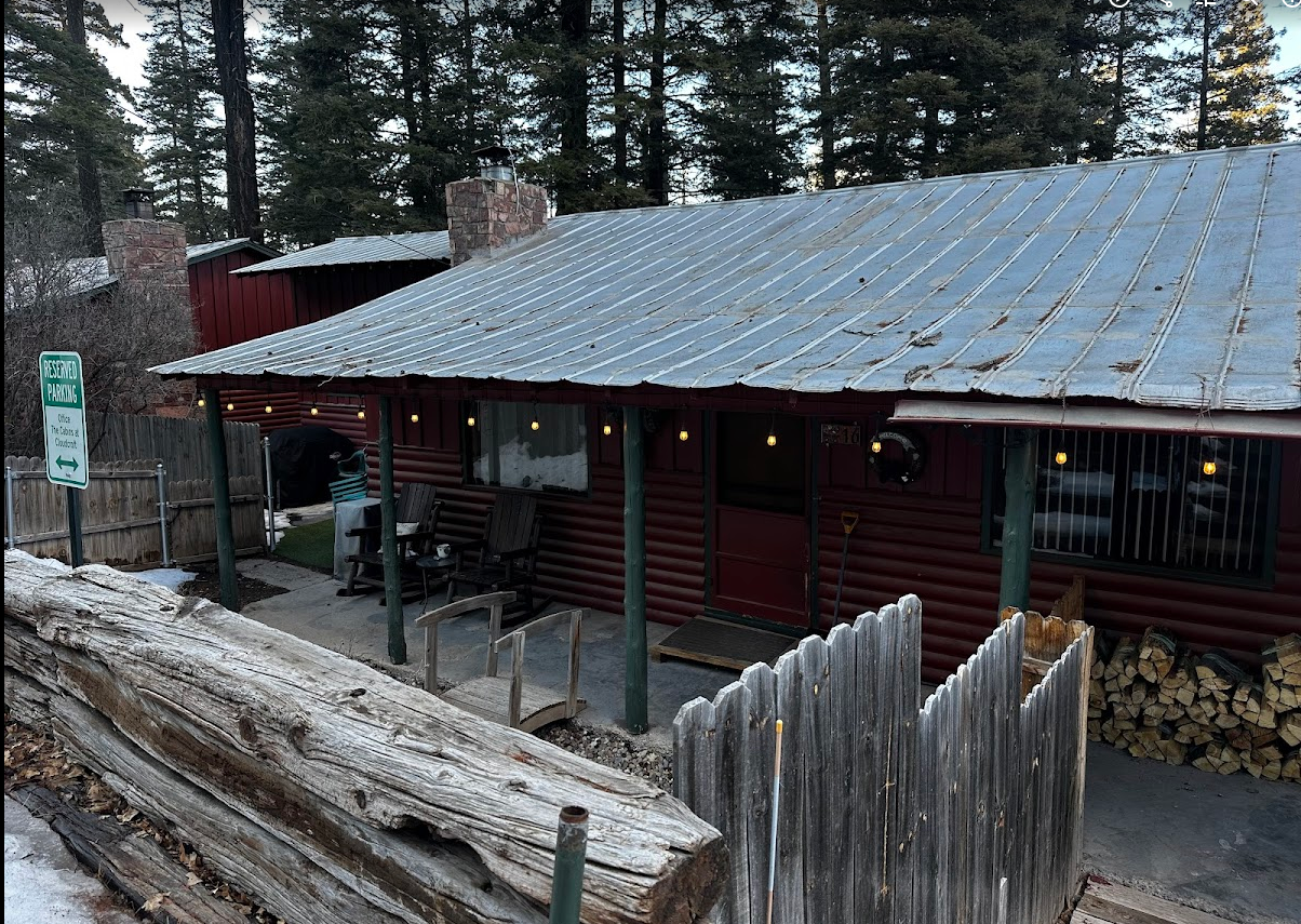 A red log cabin with a metal roof and a wood porch sits behind a rustic wooden fence in a snowy, wooded setting.