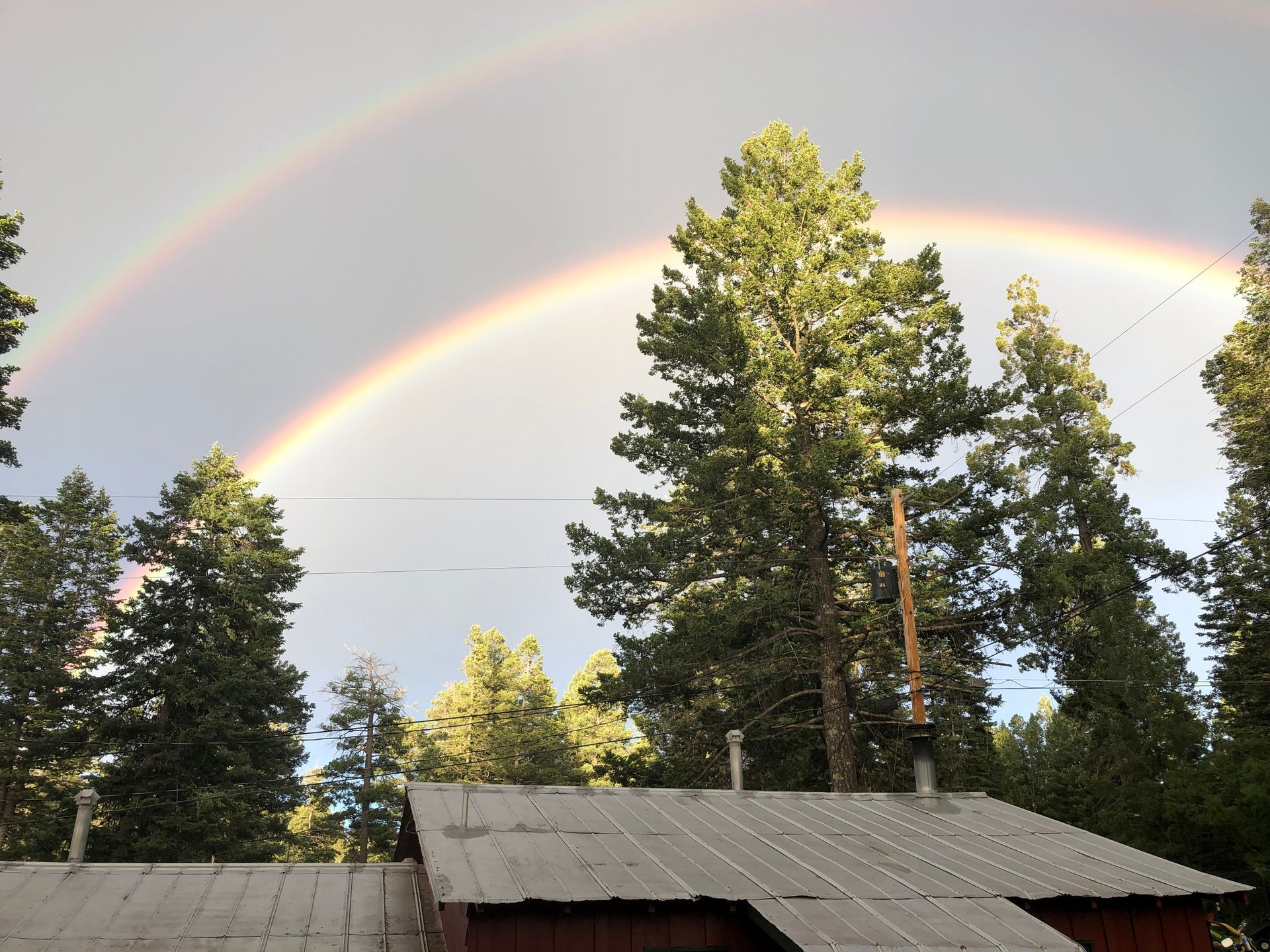 A double rainbow arches over the metal roof of a cabin surrounded by pine trees under a cloudy sky.