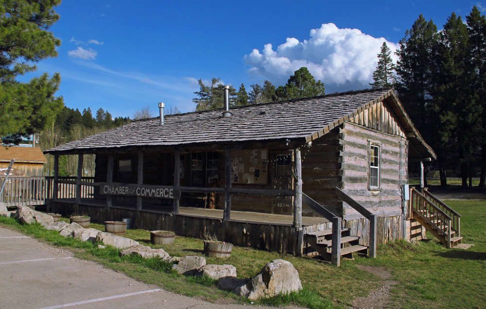 A rustic, one-story log cabin with a covered front porch, wooden shingles, and stone foundation under a blue sky.