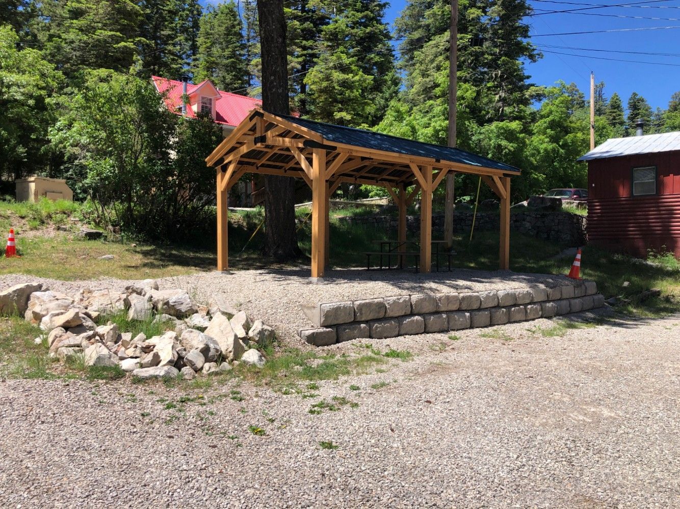 A wooden timber-frame pavilion with a black roof sits on a gravel lot next to a small red building in a wooded area.