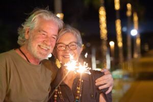 Two smiling people pose outdoors at night, holding a lit sparkler together.
