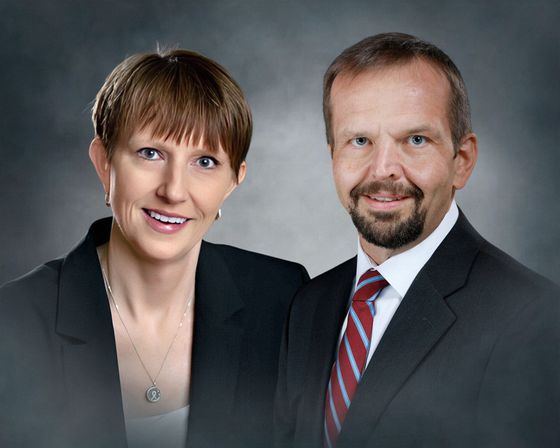 Two professionally dressed people posing against a gray studio backdrop, both smiling.