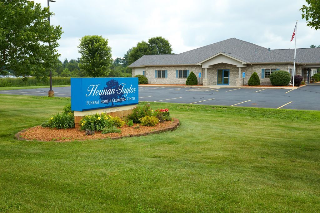 Blue sign in a landscaped lawn outside a beige building with a flagpole and parking lot