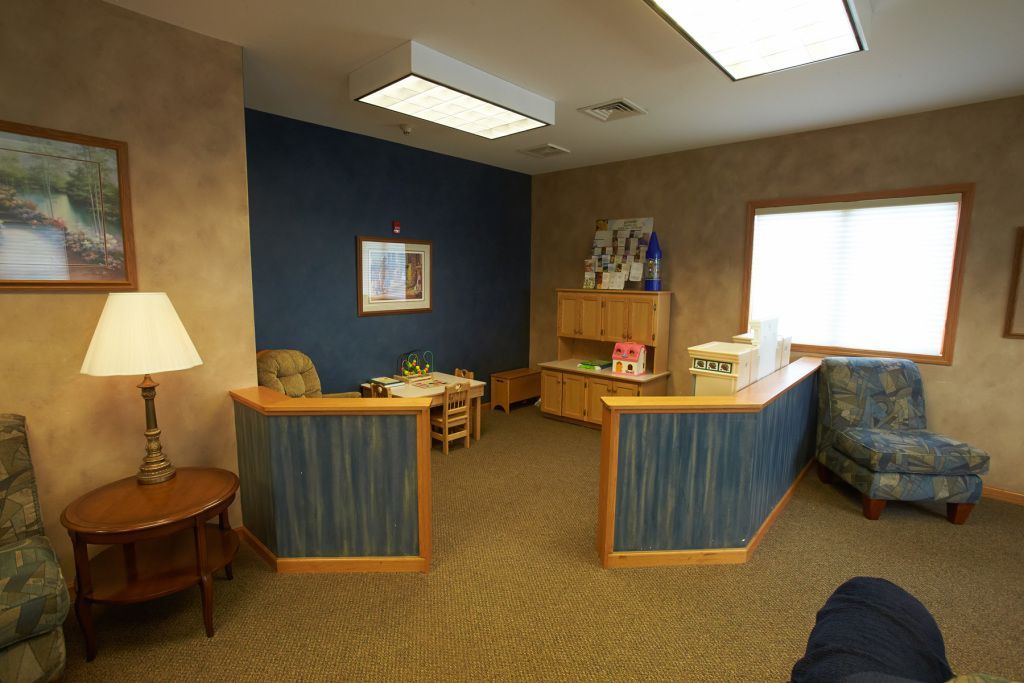 Small office waiting room with desks, blue chairs, lamp, bookshelf, and a window.