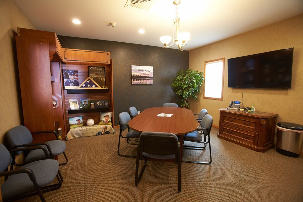 Conference room with oval table, blue chairs, wall-mounted TV, cabinet, and potted plant on carpeted floor.