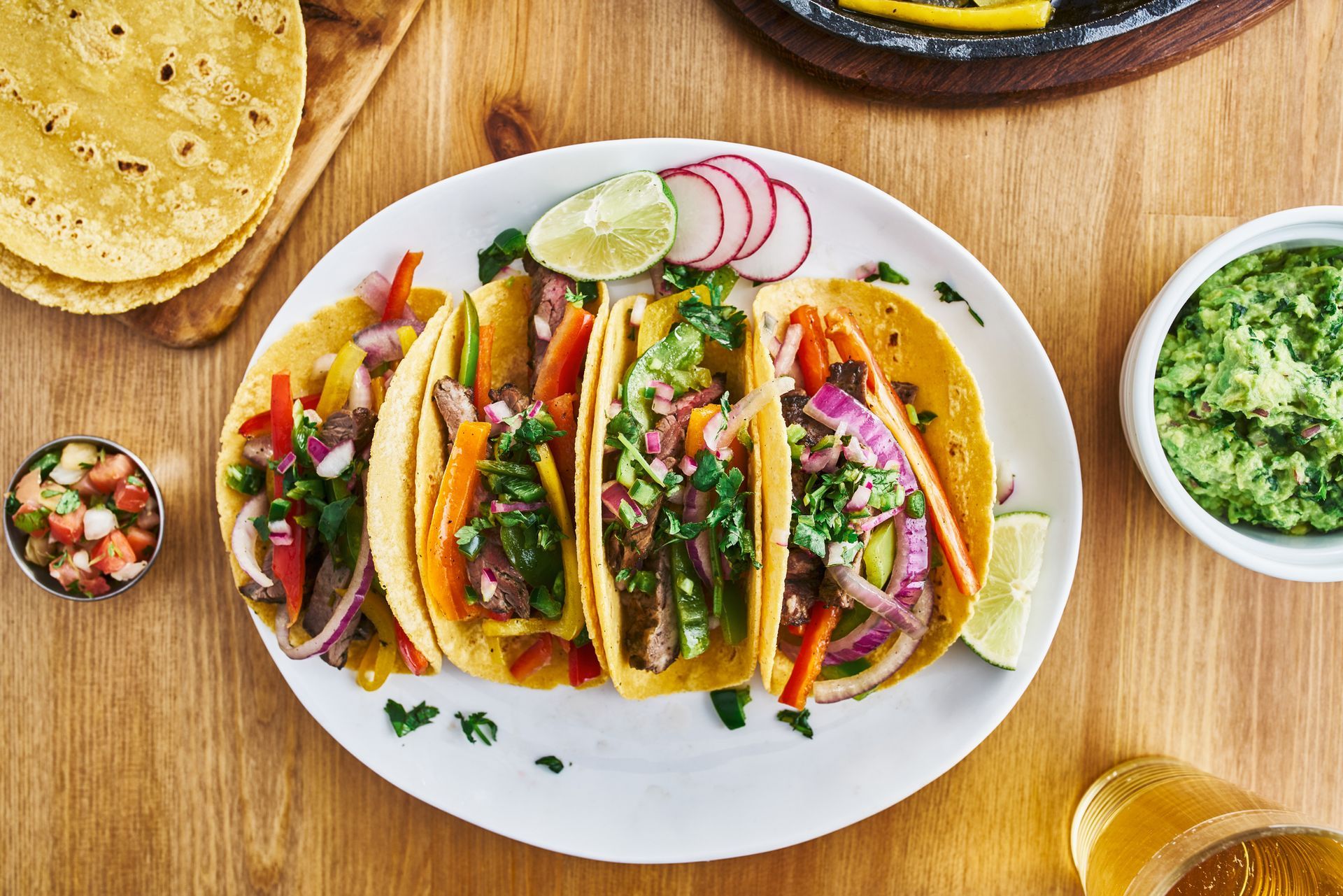 Tacos on a white plate with salsa, guacamole, radishes, lime slices, and tortillas on a wooden table.