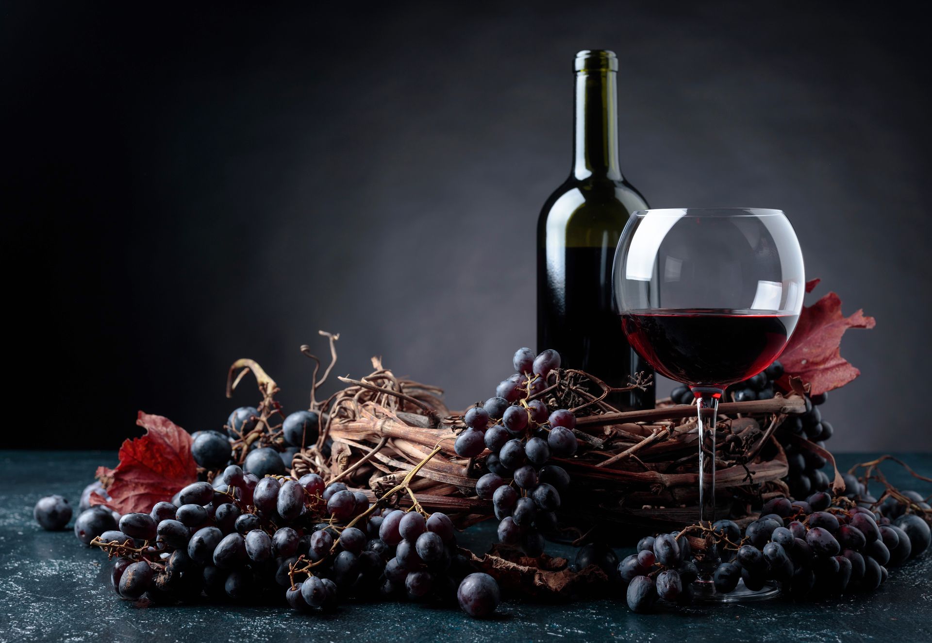 Bottle of red wine, full glass, grapes, and vine on dark background.