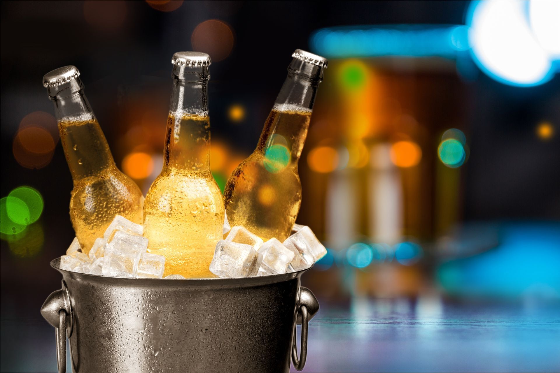 Three beer bottles in a metal bucket filled with ice cubes, in a bar setting.