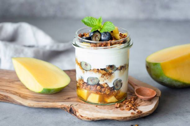 Yogurt parfait with mango, blueberries, granola, and mint in a glass jar, next to mango slices.