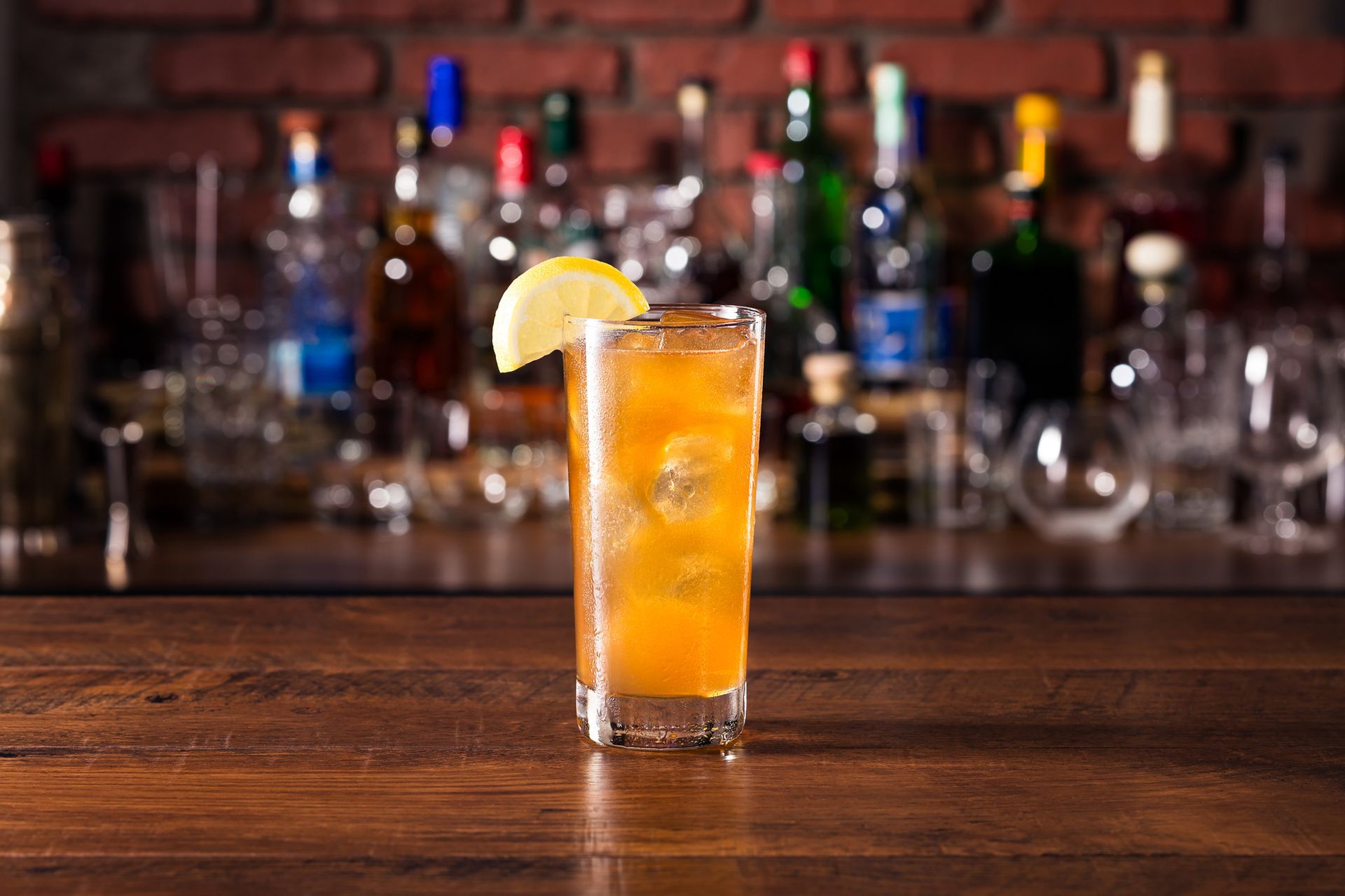 Tall glass of amber cocktail with lemon garnish on a wooden bar in front of liquor bottles.