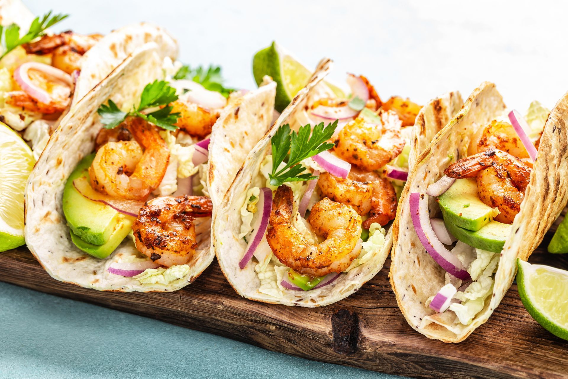 Grilled shrimp tacos with avocado, red onion, and lime wedges on a wooden board.