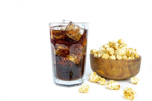Glass of cola with ice cubes and a wooden bowl of caramel popcorn.