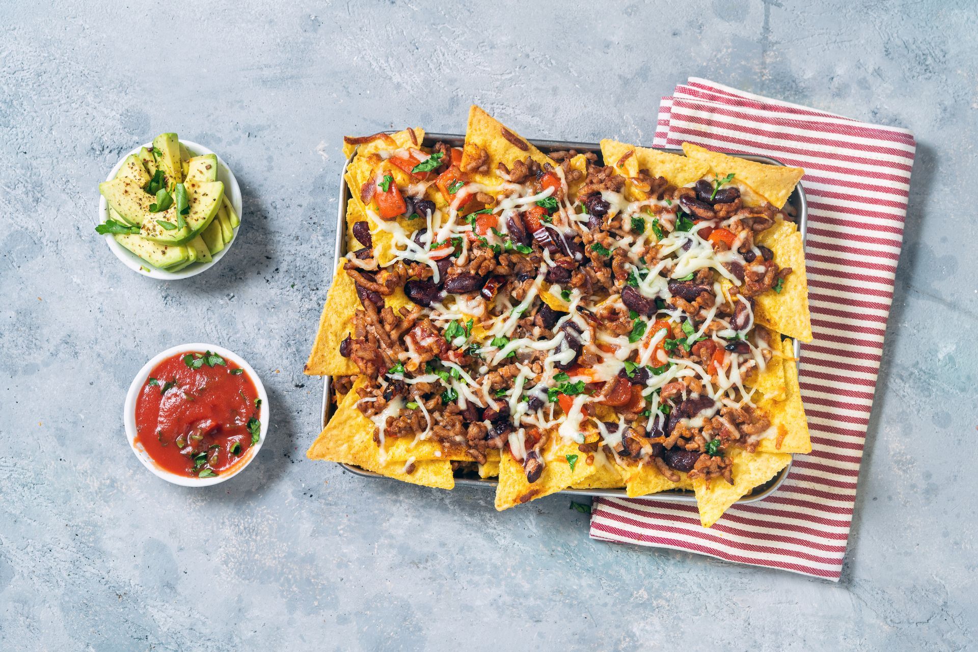 Nachos on a tray topped with cheese, olives, and meat, with salsa and guacamole.
