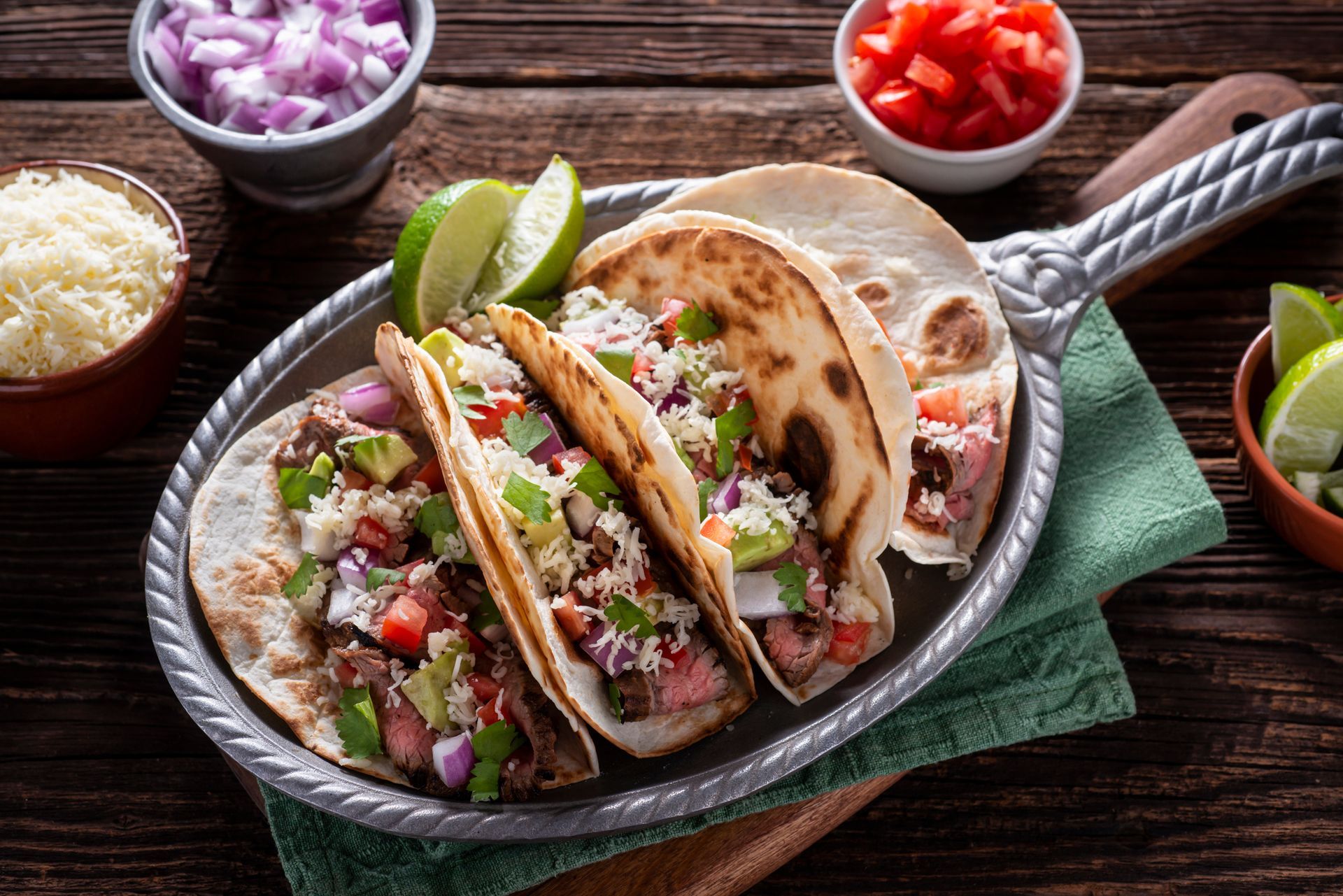 Tacos on a metal platter with toppings: tomatoes, onions, cheese, and limes on a wooden table. Tacos on a metal platter with toppings: tomatoes, onions, cheese, and limes on a wooden table.