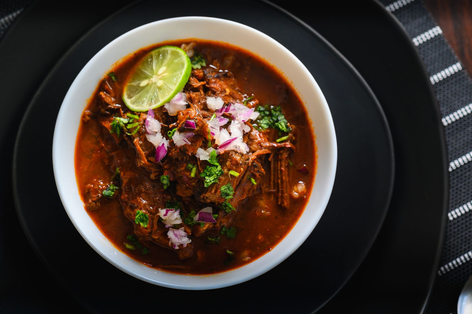 Bowl of braised meat stew with lime, onion, and cilantro garnish. Bowl of braised meat stew with lime, onion, and cilantro garnish.