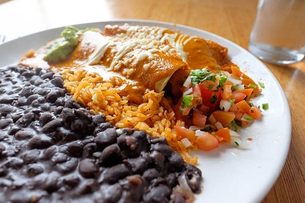 Plate of enchiladas, black beans, and rice with salsa and a glass of water on a table.