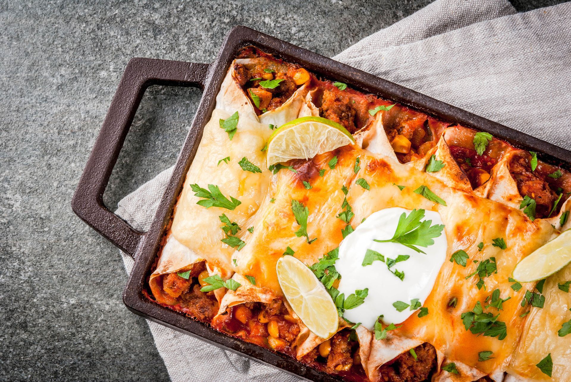 Baked enchiladas in a dark rectangular dish, topped with cheese, sour cream, lime, and cilantro, on a gray surface.