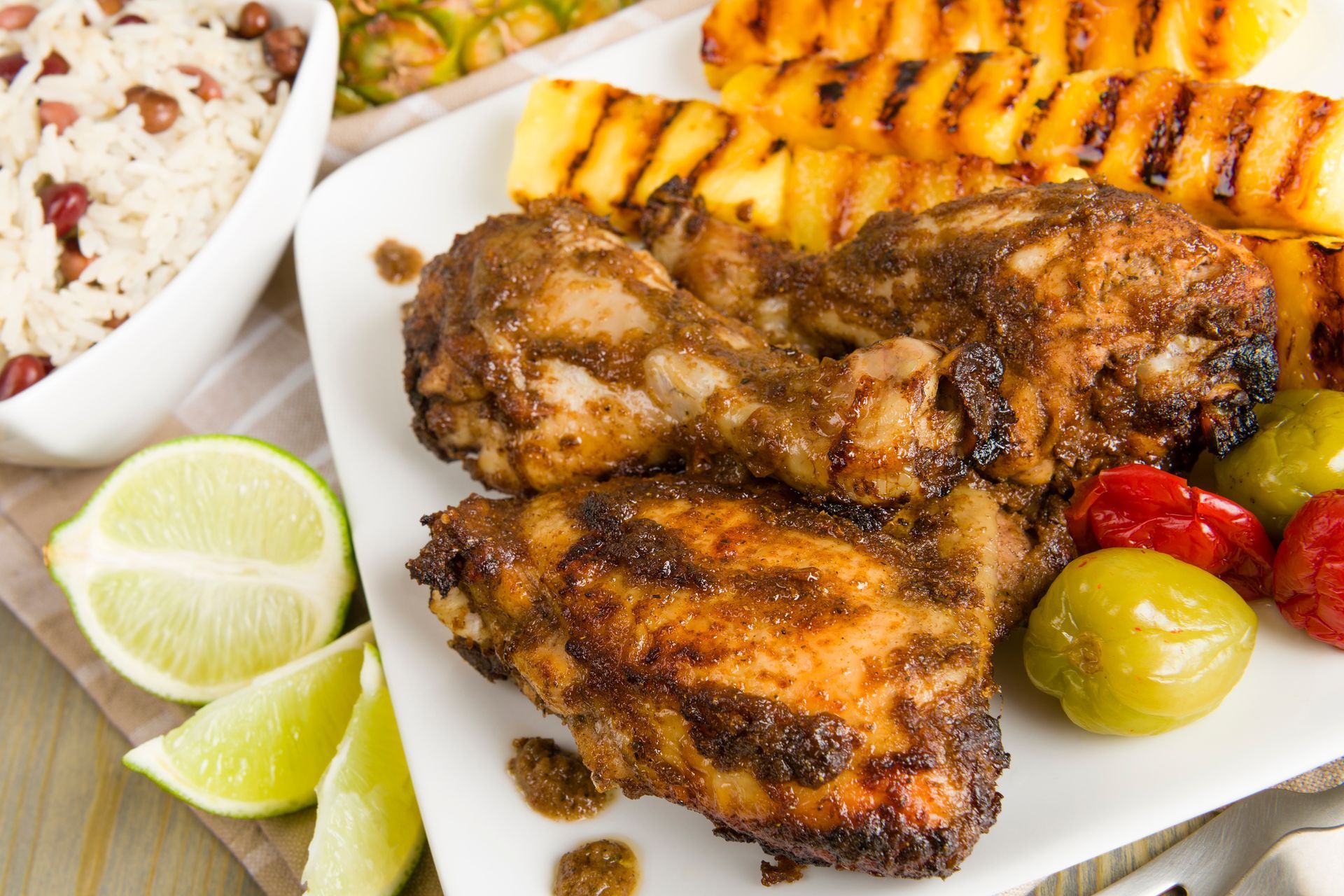 Jerk chicken with grilled pineapple, rice and peas, limes, and peppers on a white plate.