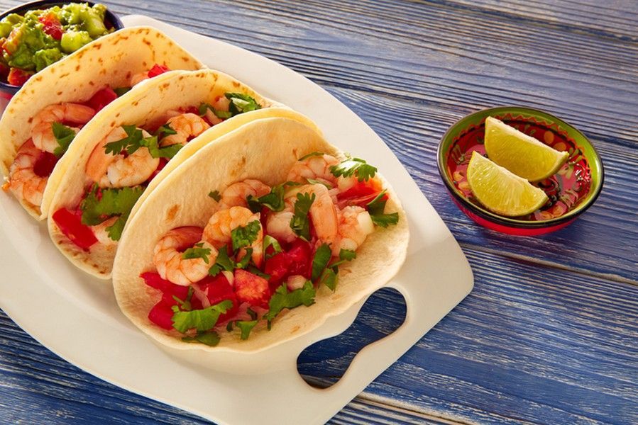 Three shrimp tacos on a white plate with salsa, guacamole, and lime wedges on a blue wooden surface.