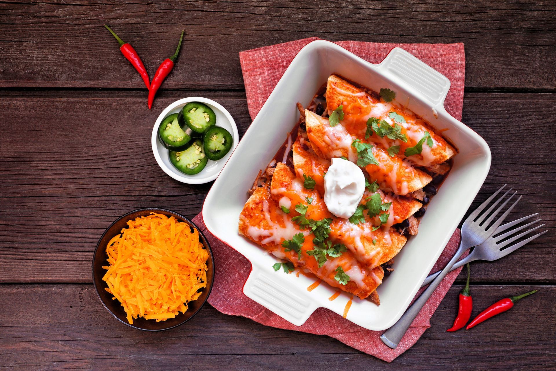 Enchiladas in a white dish, topped with sour cream and cilantro, next to jalapenos, shredded cheese, and chili peppers.