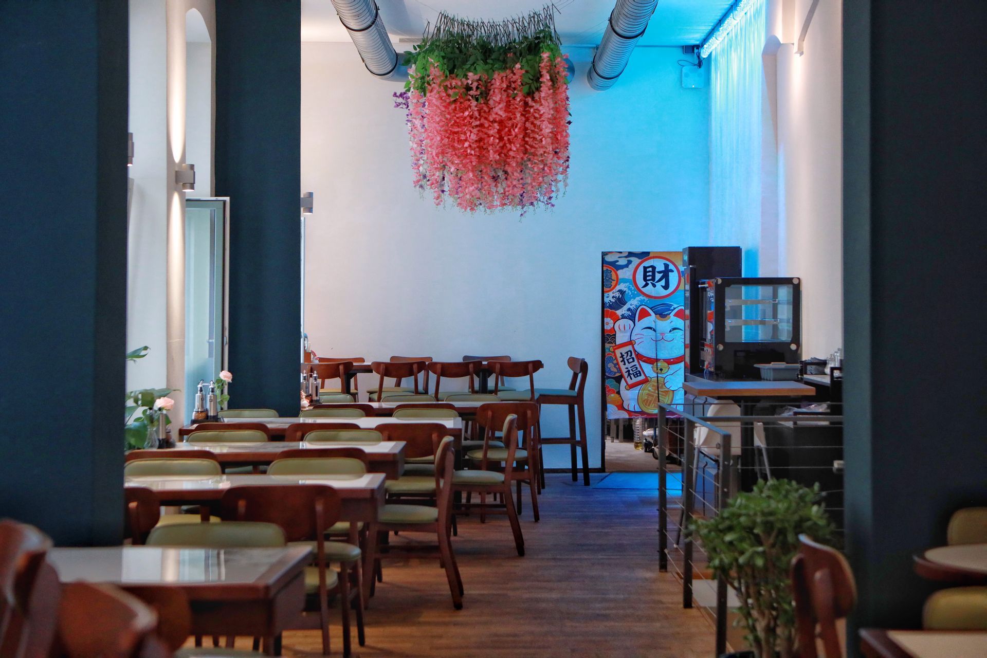 Leerer Café-Gastraum mit Holztischen, Stühlen und hängender rot-grüner Dekoration unter einer blau beleuchteten Decke
