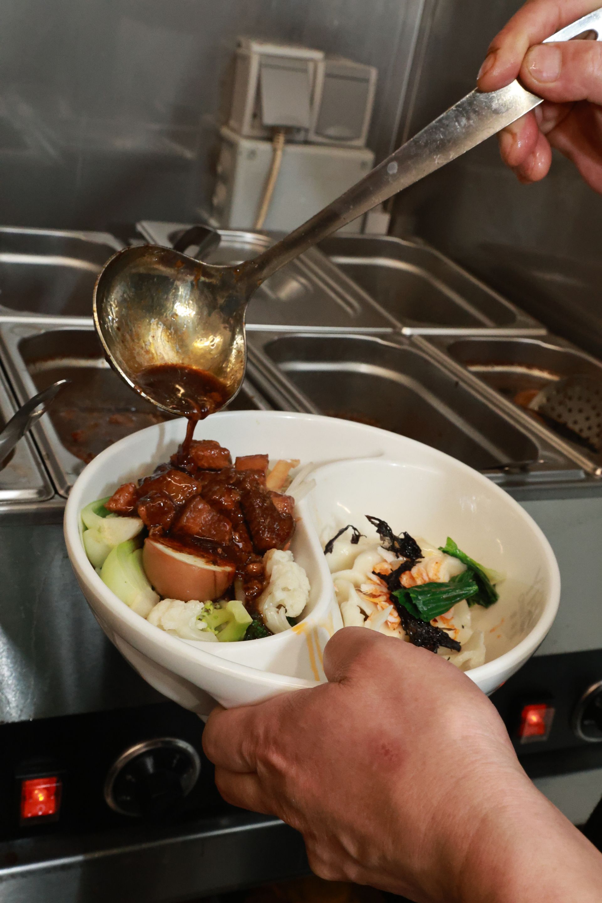 Essen wird in einer Küche mit einer Schöpfkelle in eine weiße Schüssel gefüllt; darin befinden sich Fleisch und Gemüse.